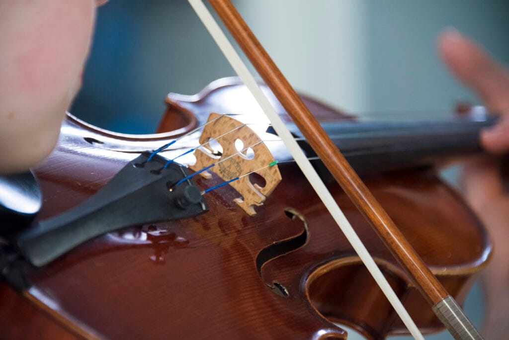 A tight shot of a student playing the violin from over the shoulder, only the bridge and the bowstrings in focus